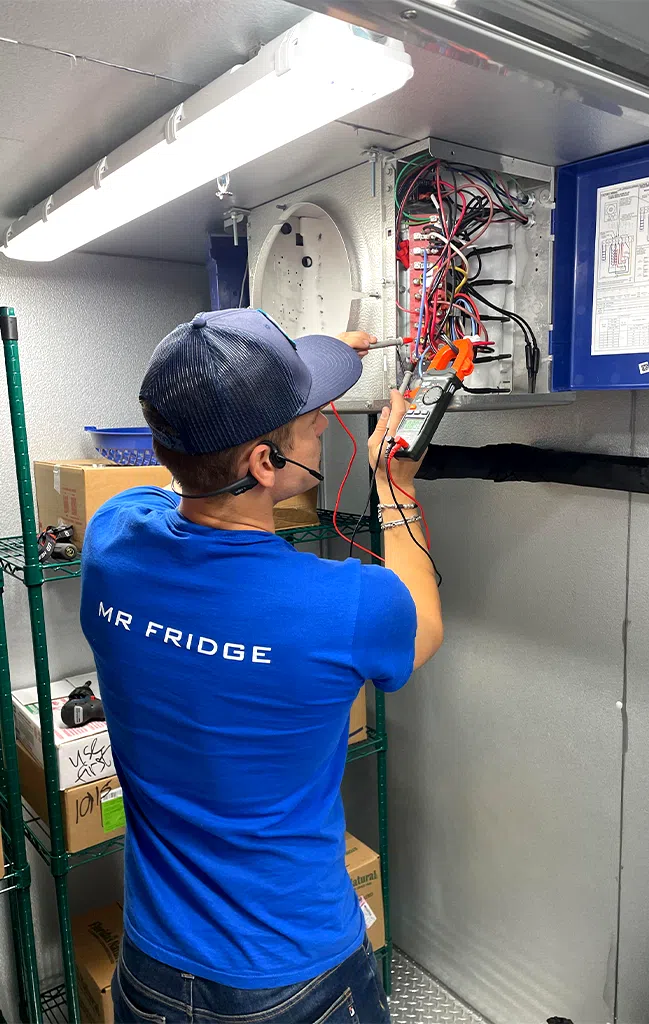 Mr. Fridge technician in a blue logo shirt repairing a commercial refrigerator inside a restaurant kitchen.